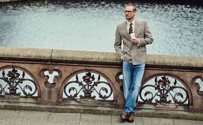 Man in glasses wearing a checked blazer, shirt and tie with blue jeans Rick, leaning against a stone railing by the water and holding a cup.