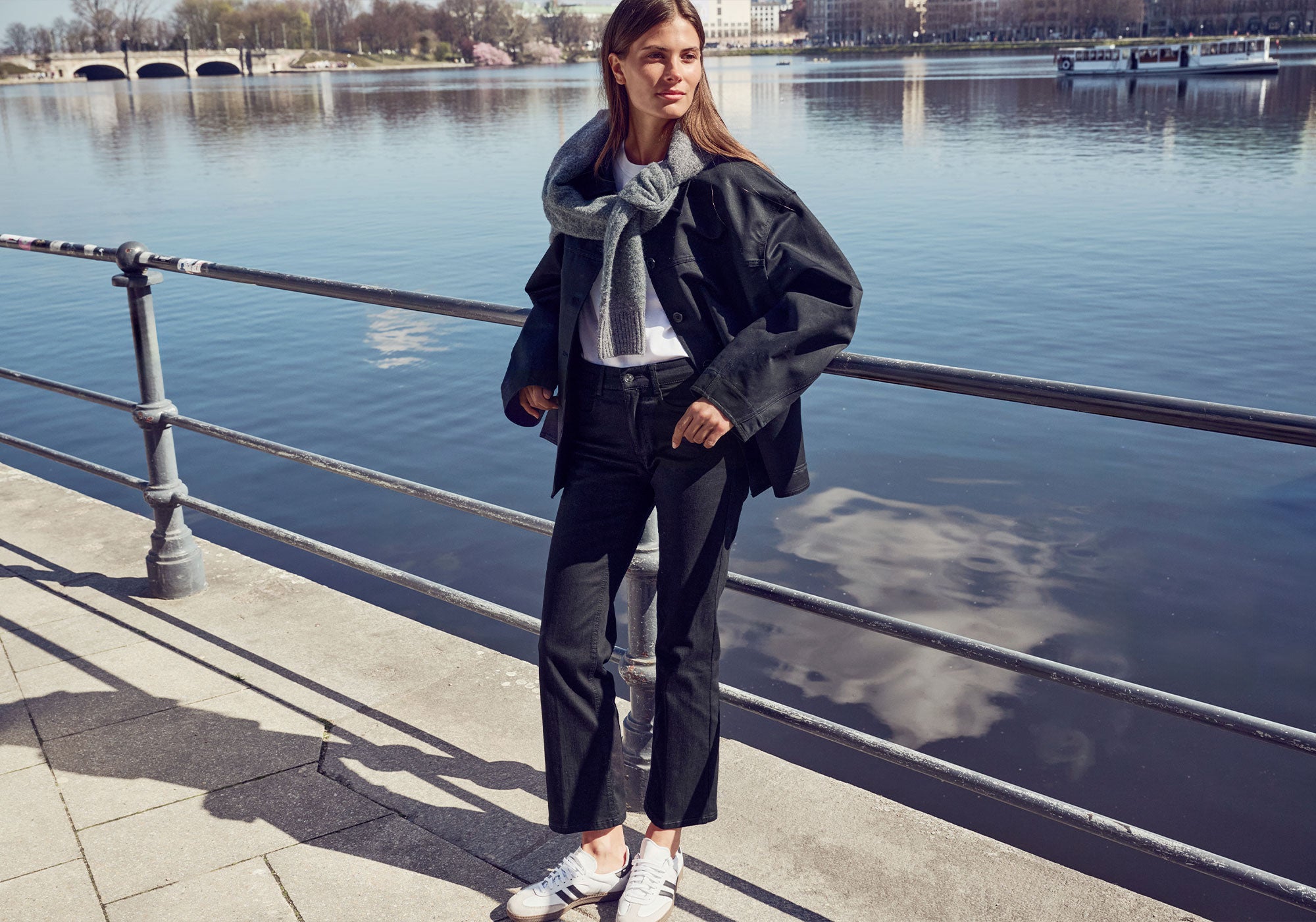 Women's model wearing a black jacket, grey knit scarf and dark wide-leg jeans by the waterfront.