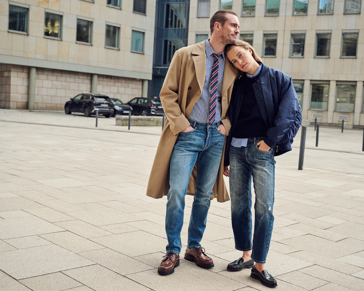 Man in a beige coat and blue jeans standing next to a woman in a navy bomber jacket and blue jeans, leaning her head on his shoulder.