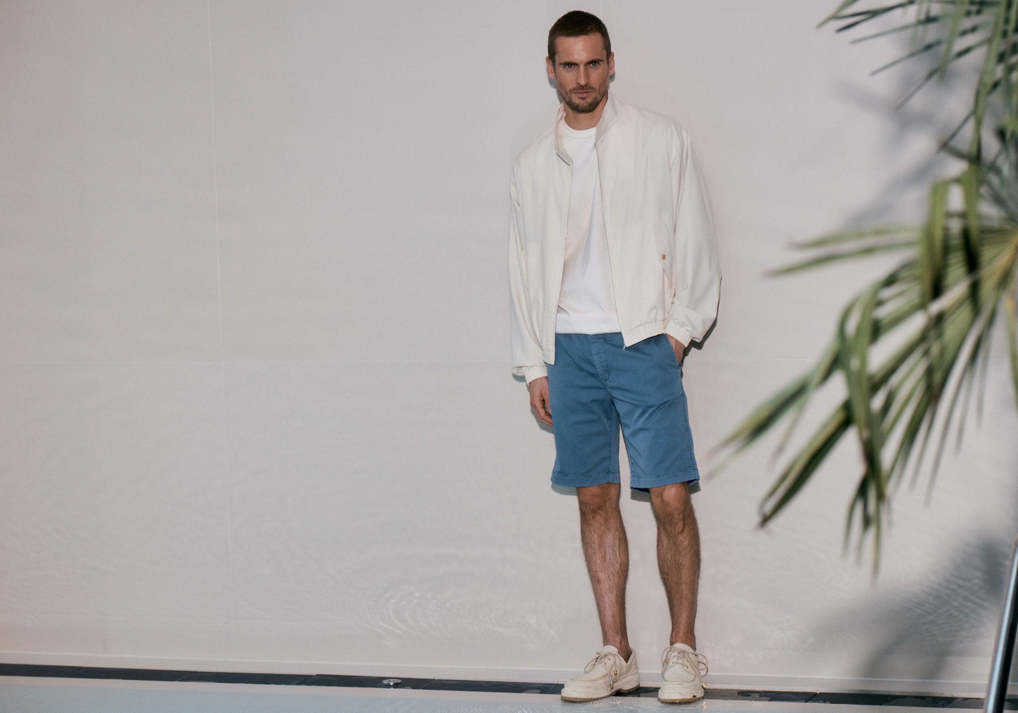 A man stands casually against a light-colored wall, wearing a white jacket over a white T-shirt, blue shorts, and light-colored sneakers. One hand is in his pocket, and he looks directly at the camera with a neutral, slightly serious expression. A blurred green plant partially frames the right side of the image, adding a soft, natural element to the otherwise minimal indoor setting.