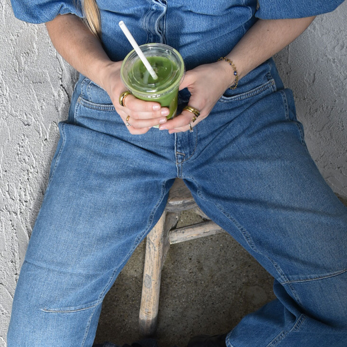 Person wearing blue denim jeans sitting on a stool while holding a green iced matcha drink.