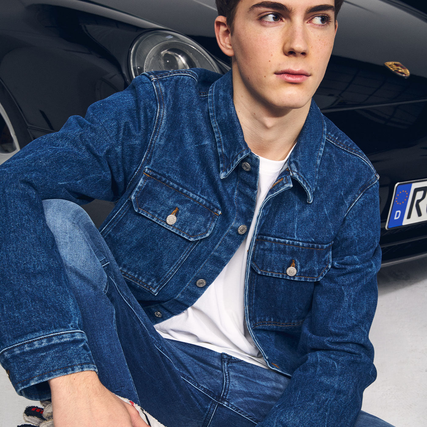 Young man wearing a blue denim jacket and jeans sits casually on the floor in front of a sports car, looking to the side in a clean studio setting.