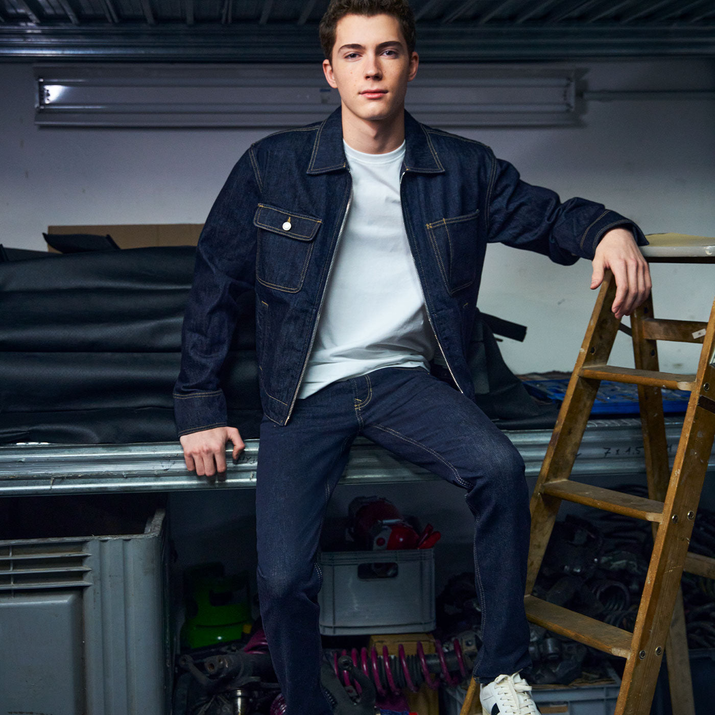 Young man wearing a dark denim jacket and jeans sits on a workbench in a garage setting, resting one arm on a wooden ladder and looking confidently at the camera.