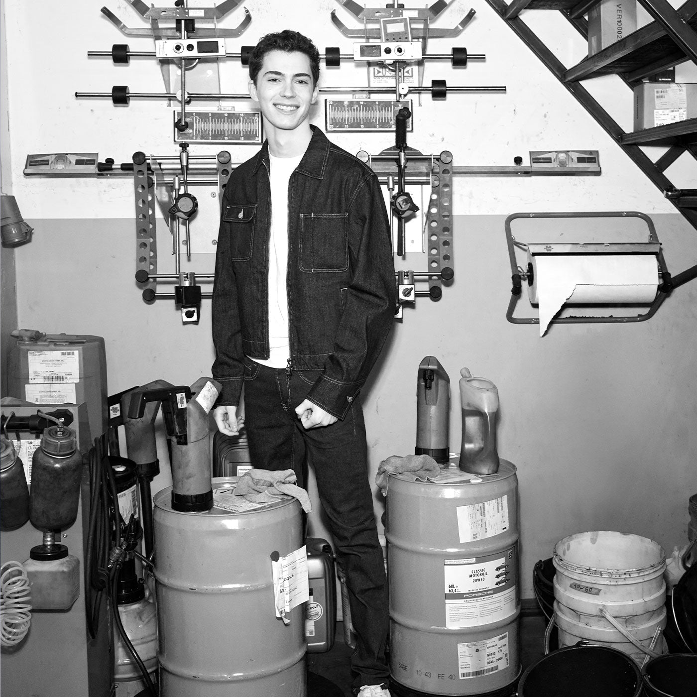 Black-and-white portrait of a young man standing in a workshop, surrounded by tools and equipment, wearing a denim jacket and jeans.