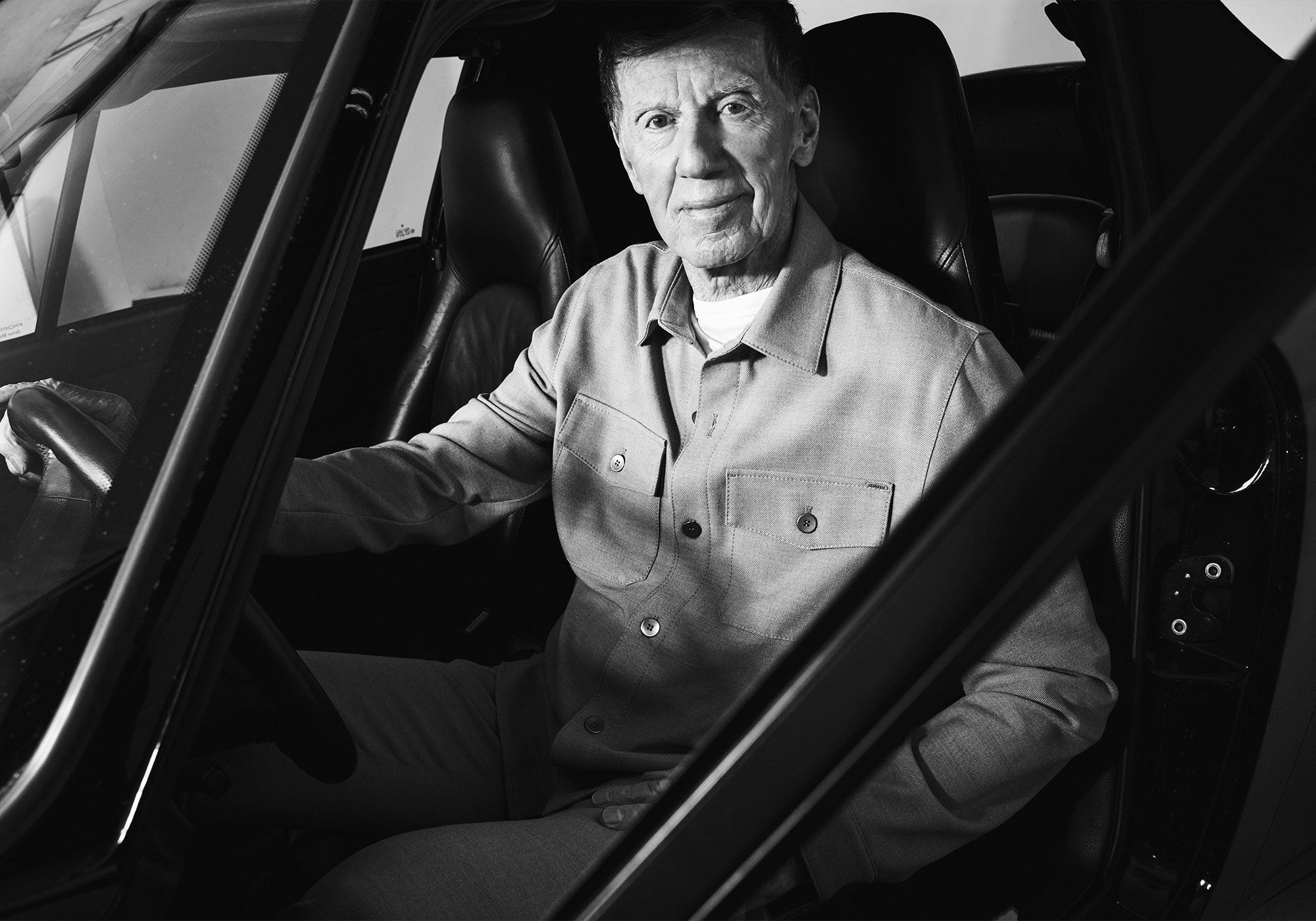 Black-and-white portrait of an older man sitting in the driver’s seat of a car, looking calmly toward the camera.