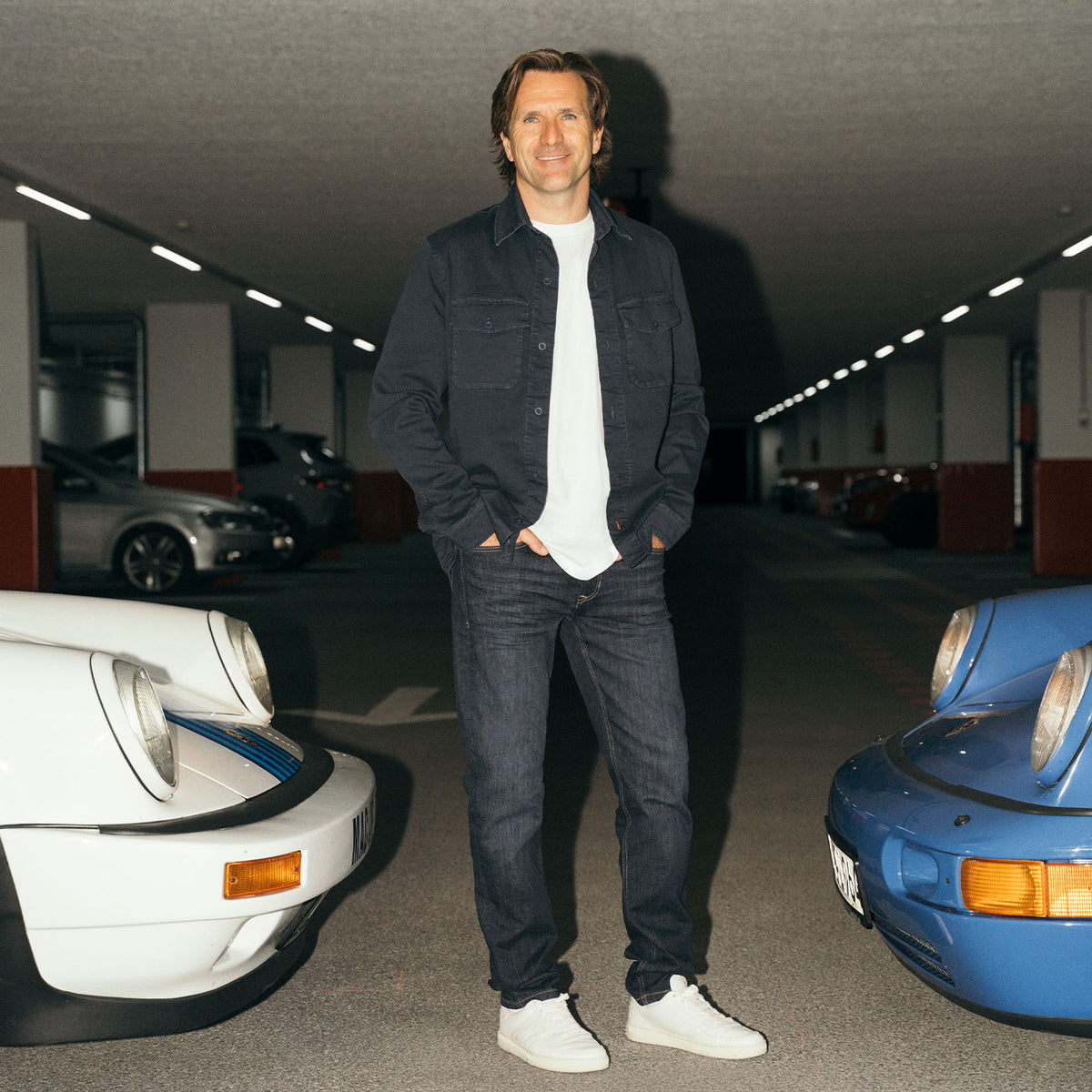 Markus Winkelhock wearing a dark MAC Jeans look, standing between two classic sports cars.
